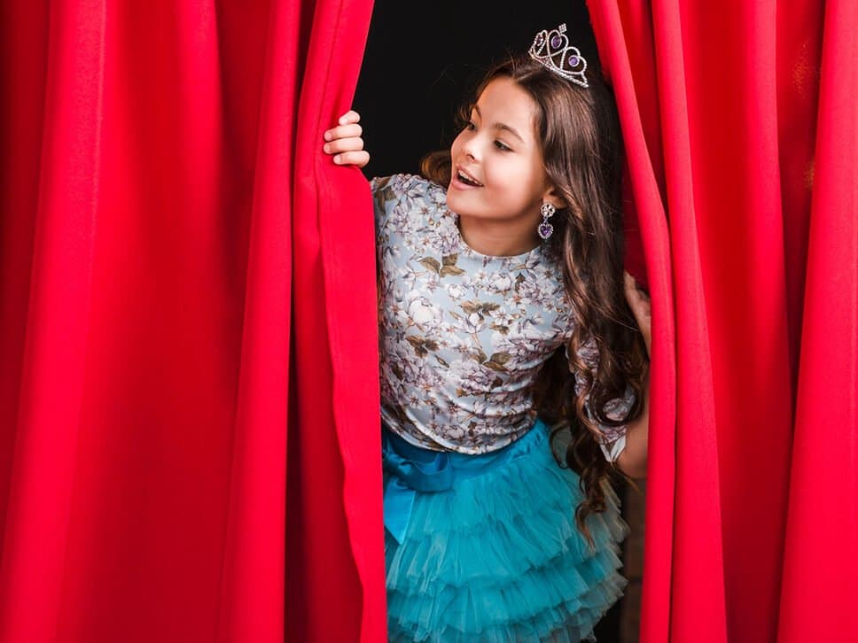 A young girl in a tiara peeks out from behind a red stage curtain.