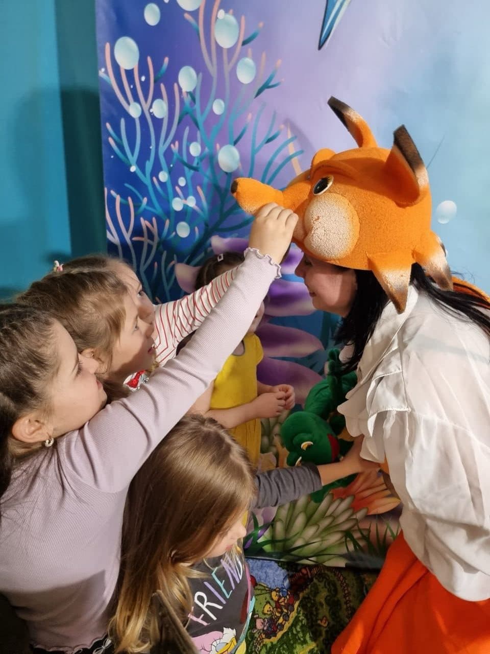 A performer in a bright fish costume leans toward excited children during the show.