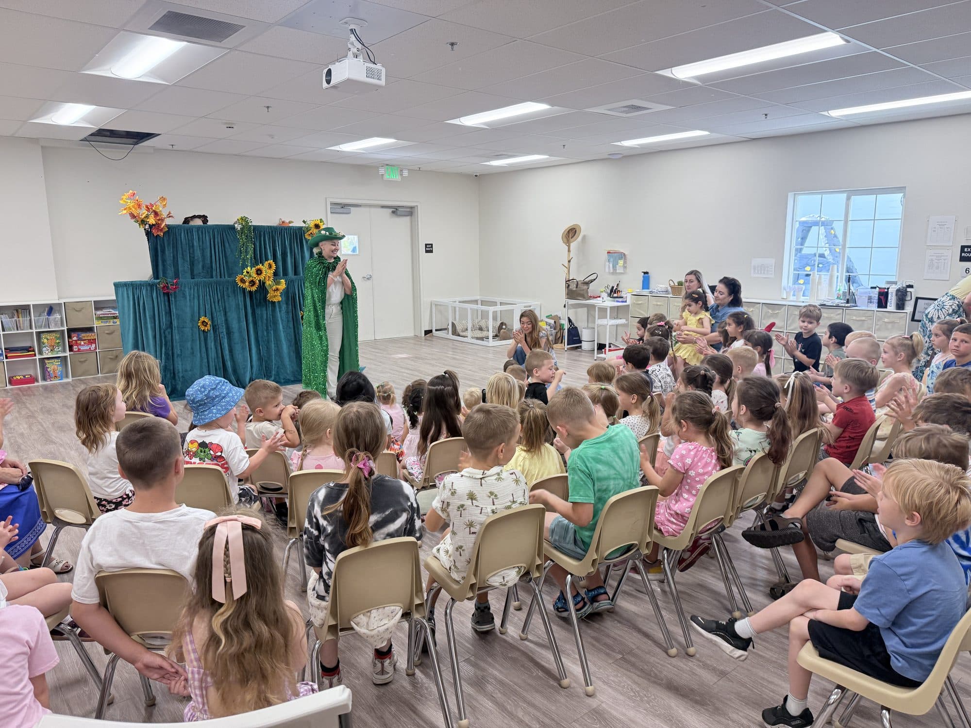 A costumed performer presents on a small stage to a large seated audience of children.