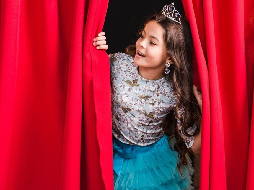 A young girl in a tiara peeks out from behind a red stage curtain.